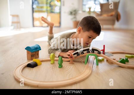 Sorridente ragazzino europeo in pigiama giace sul pavimento, gioca con la strada e le auto da sole nella stanza dei bambini Foto Stock