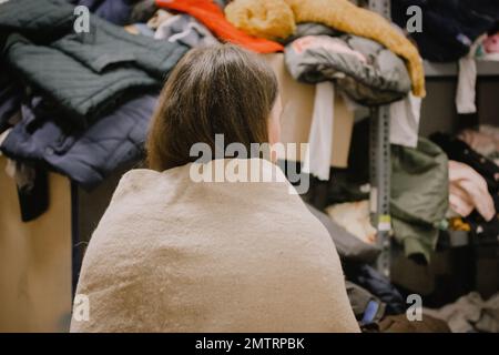 Una donna rifugiata povera senza tetto ha bisogno di aiuto nel punto di distribuzione degli aiuti umanitari. Persona senza volto avvolta in lana di plaid. Una povera donna solitaria e timida dentro Foto Stock