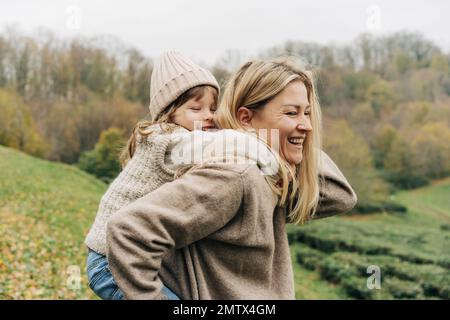 Giovane madre ridente allegra cavalca la figlia piccola sulla schiena. Foto Stock