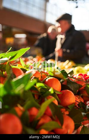Tangerini freschi su uno stallo di mercato all'interno di un mercato, con due uomini e l'interno di un mercato sfocato in background. Foto Stock
