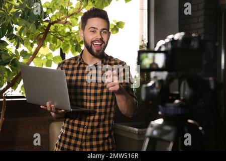 Giovane blogger con laptop che registra video su una fotocamera in interni Foto Stock