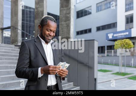 Felice afro-americano uomo, lavoratore ufficio, uomo d'affari si trova vicino al centro ufficio, tiene in mano contanti, conta soldi. Ricevuto premio finanziario, stipendio, bonus. Foto Stock