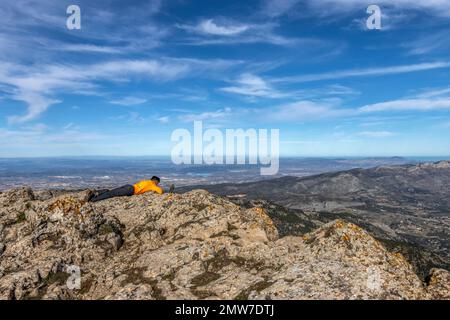 Uomo escursionista seduto sul bordo di una collina rocciosa sotto un bel cielo nuvoloso. Alpinista e un simpatico uccello ammirando la vista della valle di montagna A. Foto Stock