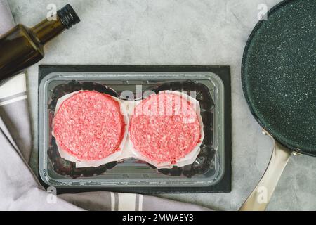 Polpettine di hamburger in plastica, olio d'oliva e padella da friggere primo piano. Hamburger freschi e crudi primo piano su uno sfondo di pietra grigia, piatto con c Foto Stock