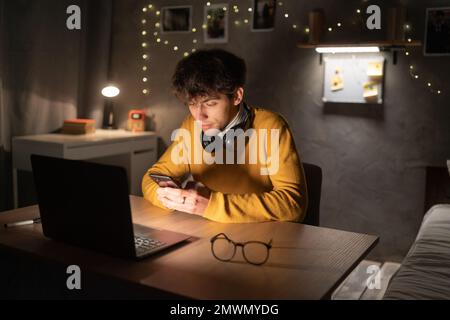 Studente giovane ragazzo che studia nel dormitorio dell'università, seduto alla scrivania, utilizzando un computer portatile, tenendo un telefono cellulare. Spazio di copia Foto Stock