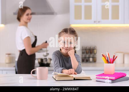 Bambina che fa i compiti mentre la madre cucina in cucina Foto Stock