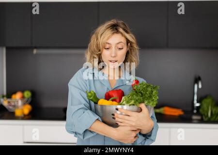 bella giovane donna con ciotola di tenuta dei capelli ondulata con verdure biologiche in cucina, immagine di scorta Foto Stock