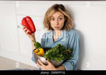 scioccato giovane donna con capelli ondulati ciotola di tenuta con verdure biologiche in cucina, immagine di scorta Foto Stock