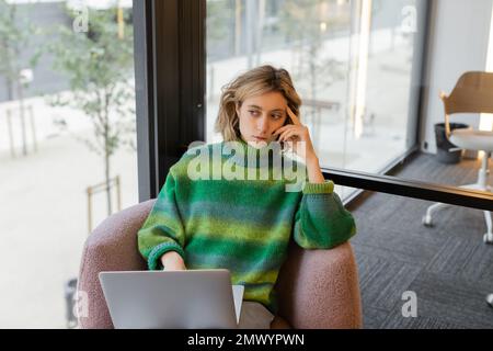 Giovane donna premurosa in maglione seduto con un computer portatile nella hall dell'hotel a Barcellona, immagine stock Foto Stock