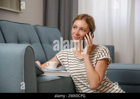 Giovane donna rilassata godendo di una giornata rilassante a casa con telefono, tazza di caffè e libro Foto Stock