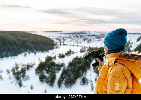 Giovane scienziata ecologista con binocolo che guarda la migrazione degli uccelli in inverno contro la foresta innevata Bird watching Wildlife manager copia spazio Foto Stock