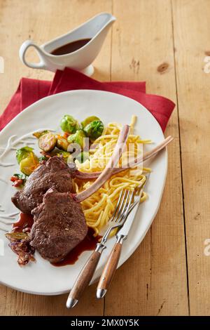 Alto angolo di appetitose bistecche di cervo di tomahawk con spaghetti serviti con sprout di Bruxelles serviti su un tavolo di legno con salsa in cucina Foto Stock