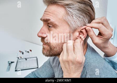 Audiologo che inserisce l'apparecchio acustico ITE nell'orecchio dell'uomo adulto presso il centro audiologico, primo piano, vista laterale. Trattamento sordità, soluzioni acustiche Foto Stock
