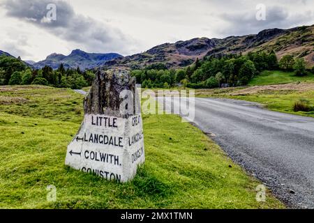 Lake District, Cumbria, Inghilterra, UK - pietra miliare in vecchio stile sulla strada fuori Elterwater villaggio con le cime di montagna dei Langdale Pikes in lontananza Foto Stock