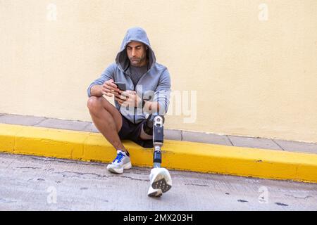 Atleta maschile di mezza età con gamba protesica seduta sul bordo e navigazione sul cellulare durante la pausa di allenamento in città. Foto Stock