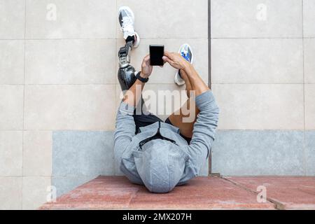 Vista dall'alto dell'atleta maschile in felpa grigia con cappuccio con gamba protesica seduta sul marciapiede vicino alla parete e guardare video sul cellulare durante la pausa durante il lavoro di fitness Foto Stock