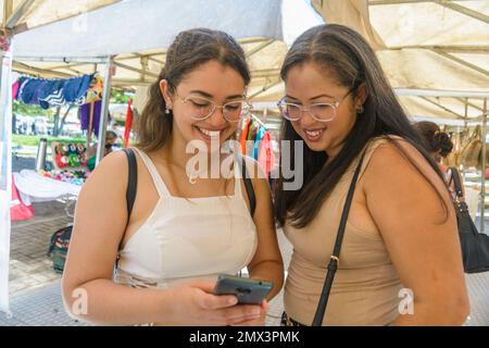Due donne adulte turisti, madre e figlia, stanno ridendo, in piedi alla fiera dell'artigianato, controllando il loro conto bancario al telefono, mentre a piedi aro Foto Stock