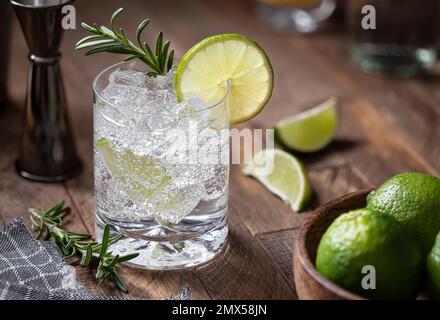 Cocktail tonico e gin con fettine di lime, rosmarino e ghiaccio su un tavolo rustico in legno Foto Stock