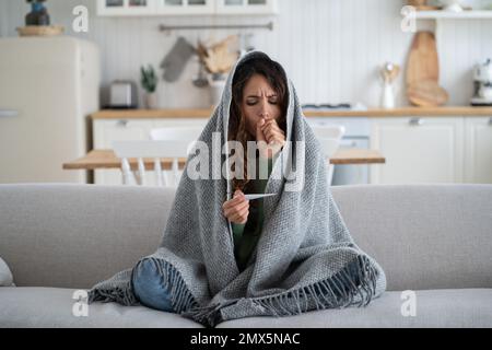 Donna malsana spaventata che guarda il termometro si siede avvolto in una calda plaid sul divano a casa Foto Stock