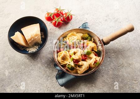 Vista dall'alto di appetitose tortelloni fatte in casa con broccoli e pomodoro vicino a una ciotola di parmigiano su un tavolo in pietra grigia Foto Stock