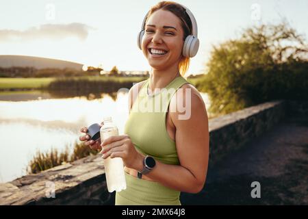 Felice sportivo che fa una pausa in acqua all'aperto. Adatta la donna in piedi accanto a un lago e guarda la fotocamera. Donna che si sta allenando con le cuffie nel Foto Stock