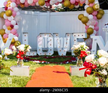 Segno d'amore dietro petali di rosa a forma di cuore con un tappeto rosso e palloncini allestiti per una proposta di marraige in un cortile residenziale. Foto Stock