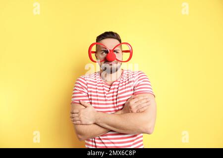 Giovane emotivo con occhiali da festa e naso di clown su sfondo giallo. Aprile Fool's Day Foto Stock