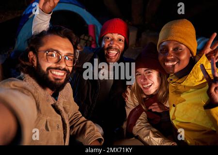 POV dell'uomo che prende un selfie con il gruppo vario degli amici, all'aperto durante il viaggio del campeggio alla notte. Foto Stock