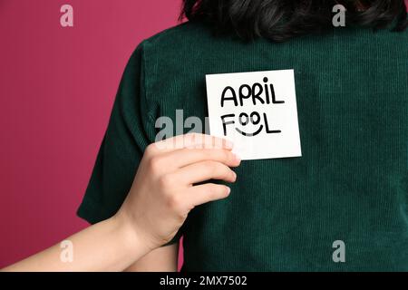 Donna che si attacca APRILE NOTA SCIOCCO alla schiena di un amico su sfondo rosa, primo piano Foto Stock