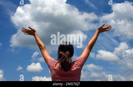 Giovane ragazza adolescente si alza, guardando verso l'alto, con le braccia allungate abbracciando il cielo. Il cielo è blu con grandi nuvole soffici. Foto Stock