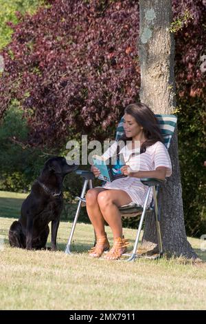 giovane donna seduta in giardino leggendo un libro con labrador nero Foto Stock