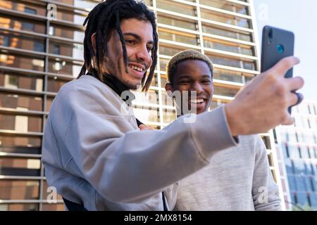 Amici maschi multirazziali positivi in abiti casual sorridendo e guardando lo schermo dello smartphone mentre si prende selfie contro l'edificio moderno Foto Stock