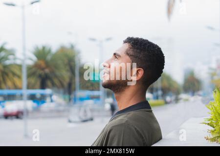 Vista laterale dell'etnia latina maschile in stile casual che si affaccia su un muro sulla strada contro un edificio moderno Foto Stock