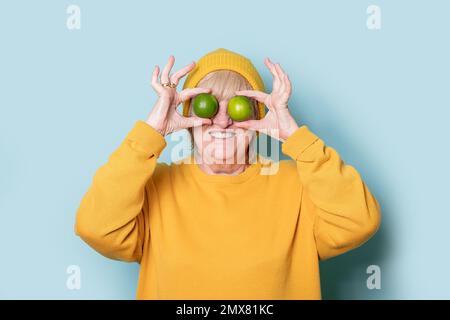 Donna anziana allegra in maglione giallo e cappello che copre gli occhi con lime su sfondo blu Foto Stock