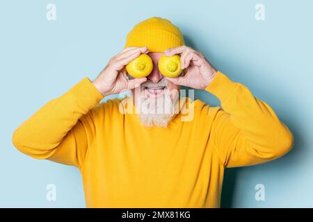 Sorpreso uomo anziano bearded in giallo maglione e cappello che copre gli occhi con limoni su sfondo blu Foto Stock