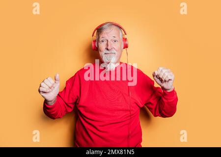 Hipster maschile senior content con barba in camicia rossa ascoltando musica in cuffie e ballando mentre si guarda la fotocamera su sfondo giallo Foto Stock