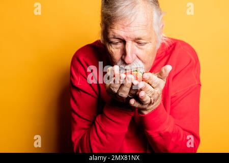 Crop calmo uomo anziano in camicia rossa odorante fragola aromatica in piedi su sfondo giallo Foto Stock