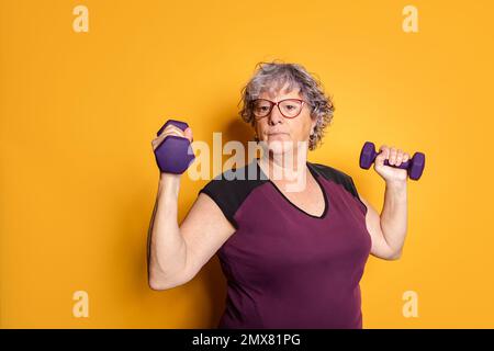 Potente donna anziana in occhiali e abbigliamento sportivo che solleva manubri mentre si allenano su sfondo giallo Foto Stock