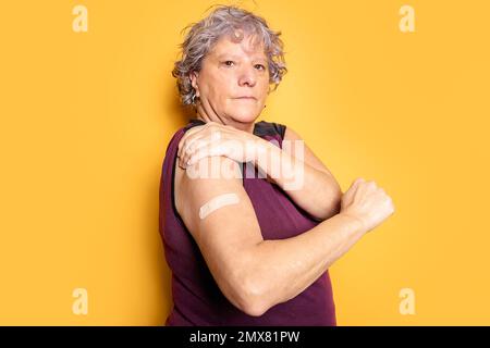 Donna senior seria in t shirt guardando la fotocamera, mentre dimostra la patch medica su sfondo giallo Foto Stock