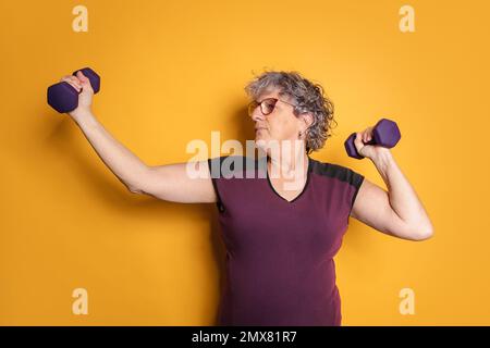 Potente donna anziana in occhiali e abbigliamento sportivo che solleva manubri mentre si allenano su sfondo giallo Foto Stock