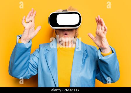 Donna anziana stupita in abiti luminosi che sperimentano la realtà virtuale con le mani sollevate mentre si trova in piedi contro lo sfondo giallo negli occhiali per la realtà virtuale Foto Stock