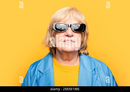 Donna anziana allegra in abiti luminosi e occhiali da sole sorridendo e guardando la macchina fotografica mentre si è in piedi su sfondo giallo Foto Stock