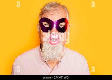 Positivo uomo anziano barbuto in camicia rosa con maschera carnevale e labbra divertenti guardando la macchina fotografica su sfondo giallo Foto Stock