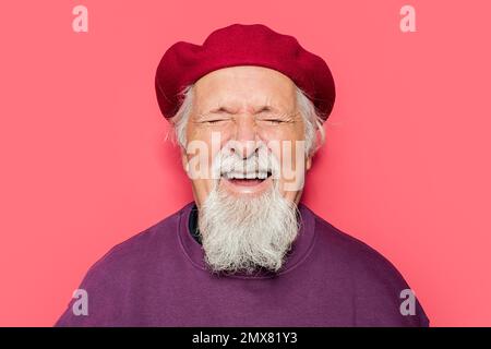Ritratto di ottimista bearded anziano maschio hipster in beret ridendo felicemente contro lo sfondo rosa con gli occhi chiusi Foto Stock