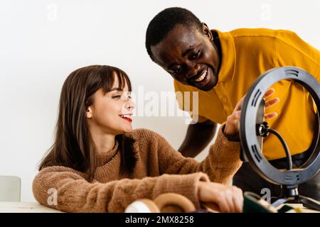 Coppia positiva giovane multirazziale in abiti casual sorridendo e guardando lo schermo dello smartphone durante la registrazione video con lampada ad anello contro wal bianco Foto Stock