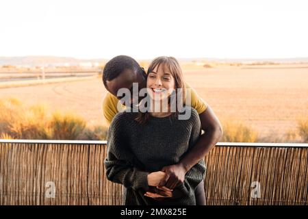 Felice giovane coppia multirazziale in abbigliamento casual sorridente e abbracciante mentre si trova in piedi sul balcone di casa e appoggiandosi sulla ringhiera contro il cielo tramonto Foto Stock