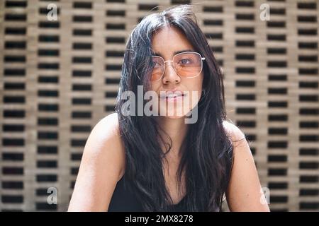 Modello femminile etnico sicuro per tutta la lunghezza in occhiali da sole con capelli lunghi guardando la macchina fotografica e pensando mentre si rilassa sulla strada Foto Stock