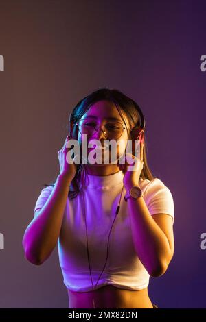 Giovane donna sorridente e sicura in un abito casual indossando cuffie e guardando lontano mentre ascolti la musica su sfondo viola Foto Stock