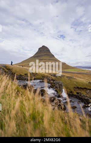 Pittoresco scenario di piccola cascata che scorre attraverso scogliera rocciosa vicino alta montagna e silhouettes turistiche e sotto cielo nuvoloso Foto Stock