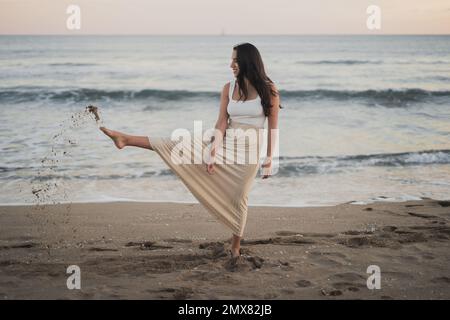 Tutta la lunghezza del soddisfare giovane ragazza etnica viaggiatore con lunghi capelli scuri in estate casual indossare sabbia calciante e sorridente mentre si trova in piedi sulla spiaggia vicino a wa Foto Stock